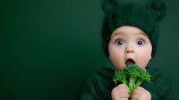 Young baby wearing green knitted hat with ears and matching sweater holds fresh broccoli floret, expressing surprise against dark green background.