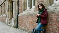Young woman in burgundy jacket and green scarf using smartphone while leaning against brick wall on urban street. Casual lifestyle and mobile technology concept.