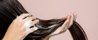 Woman applying hair treatment mask to long dark brown hair strands with hands against soft pink background for healthy haircare routine.