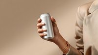 Female hand with gold jewelry holding blank aluminum can against beige background, minimal luxury product photography style for beverage marketing.