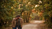 Young couple walking together on autumn forest path with golden sunlight filtering through colorful fall foliage creating romantic outdoor scene.