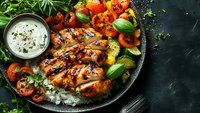 Grilled chicken breast with charred marks served on rice with fresh vegetables and creamy sauce, garnished with herbs on dark plate against black background.