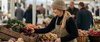 Young blonde woman in beige knit hat shopping for fresh vegetables at bustling farmers market with wooden crates full of organic produce.