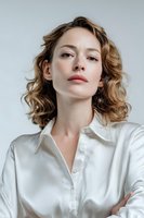 Young woman with curly brown hair wearing white silk shirt against light background, confident pose with arms crossed for fashion or beauty advertising.