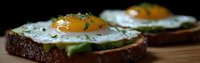 Sunny side up eggs on dark bread with sliced avocado and fresh herbs, served on wooden surface. Close up view shows rich yolks and breakfast details.
