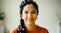 Young woman with dark braided hair adorned with white flowers, wearing rust-colored top, smiling at camera in bright indoor setting. Portrait for lifestyle, beauty, and wellness content.