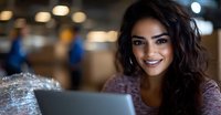 Young Hispanic woman with curly dark hair smiling while working on tablet in modern office space with blurred coworkers and warm lighting in background.