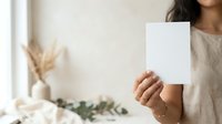 Woman of South Asian descent holding blank white card in bright interior space with dried flowers and greenery, suitable for greeting cards, invitations, and marketing mockups.
