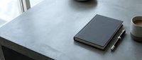 Minimalist workspace with black notebook and pen on concrete desk surface near window with natural light for productivity and planning.