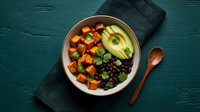 Nutritious Buddha bowl with roasted sweet potato, black beans, avocado slices, and fresh vegetables on teal wooden surface. Healthy meal prep and plant-based diet concept.