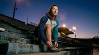 Young woman with long dark hair and fair complexion, wearing casual athletic clothing, crouching on concrete structure at dusk with urban bridge and street lights in background.