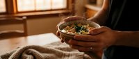 Young adult male with medium skin tone holding a warm bowl of vegetable soup or stew with fresh herbs, seated indoors near a window during golden hour lighting.