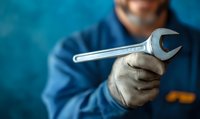 Mechanic in blue uniform holding adjustable wrench against blurred turquoise background. Close-up view of gloved hand gripping professional tool.