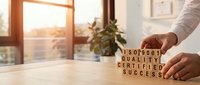 Man arranging wooden blocks spelling quality certified success on desk by window with sunset light, representing business achievement and quality assurance.