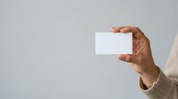 Person of Middle Eastern or South Asian descent holding blank white business card or paper against neutral gray background, suitable for corporate branding and professional mockup applications.