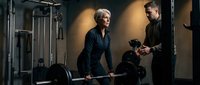Mature blonde woman exercising with barbell while male trainer provides guidance in modern gym setting with professional lighting.
