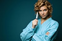 Woman with wavy blonde hair, fair complexion, wearing light blue shirt, holding kitchen knife against teal background for culinary or cooking concepts.