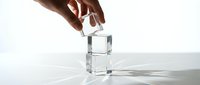 Hand carefully placing transparent glass cube on top of another creating balanced stack with dramatic shadows on white surface for minimalist design concepts.