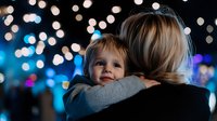 Young Caucasian child hugging parent from behind against blurred festive bokeh lights background creating magical evening atmosphere at outdoor celebration.
