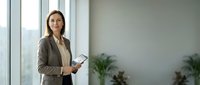Professional businesswoman holding tablet in modern office with large windows and plants creating bright workspace atmosphere.
