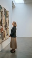 Middle-aged Caucasian woman in business attire contemplating abstract artwork in modern gallery space with natural lighting from skylight above.