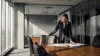 Professional businesswoman in dark suit standing confidently at conference table in modern glass office boardroom for corporate presentations and meetings.