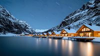 Illuminated wooden cabins reflect on calm lake waters at night, snow-covered mountains frame cozy winter resort lodges with warm glowing windows beneath star trails.