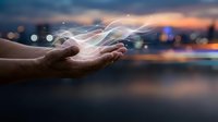 Hands cupped holding glowing digital energy streams with light particles against blurred city skyline at sunset. Technology innovation and future concepts.