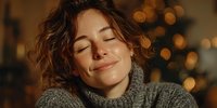 Young Caucasian woman with closed eyes and serene smile wearing gray turtleneck sweater against warm bokeh holiday lights background.