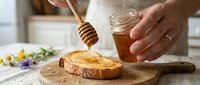 Hands drizzling honey onto toasted bread slice on wooden board with jar and flowers in background, food preparation and breakfast concept.