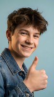 Happy teenage boy with curly brown hair giving thumbs up gesture wearing denim shirt on light blue background for youth lifestyle and positivity concepts.