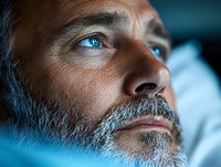 Close up portrait of mature bearded man with striking blue eyes in dramatic lighting, side view against dark background showing contemplative expression.