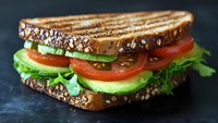 Fresh vegetable sandwich with toasted multigrain bread, layered with crisp lettuce, ripe tomato slices and creamy avocado on dark background, close up view.
