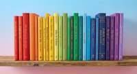 Rainbow colored books arranged in spectrum order on wooden shelf against blue wall creating vibrant library display for education and reading concepts.