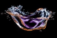 Dynamic purple liquid splash with golden edges creating crown-like formation against black background. High-speed photography captures water droplets and bubbles.