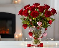 Red roses bouquet in crystal vase with heart decorations and fairy lights creating romantic Valentine's Day atmosphere by cozy fireplace.
