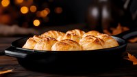 Fresh baked dinner rolls with golden brown crust in cast iron skillet on rustic wooden table with warm autumn ambiance and bokeh lights in background.