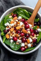 Fresh spinach salad with cranberries, almonds, and feta cheese in white bowl. Wooden spoon and dark background create appetizing healthy meal presentation.