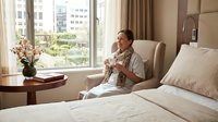 Middle-aged Caucasian woman relaxing in luxury hotel room with coffee, enjoying city view from comfortable armchair near large window with natural light.