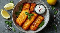 Crispy breaded fish fillets served with creamy sauce, fresh basil leaves and lemon wedges on gray ceramic plate against dark stone background. Food photography.