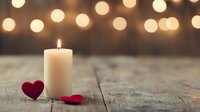 Romantic composition with glowing white candle and red felt hearts on rustic wooden surface against warm bokeh lights background, creating cozy atmosphere.