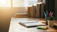 Cozy study workspace with open notebook, colorful pens, small plant and stacked books on wooden desk bathed in warm natural sunlight for productivity.