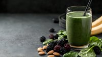 Green smoothie in glass with straw surrounded by fresh berries, almonds, and mint leaves on dark surface for healthy beverage and nutrition content.