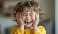 Joyful child in yellow t-shirt laughing with closed eyes and hands on cheeks, expressing pure happiness and genuine emotion in natural light setting.