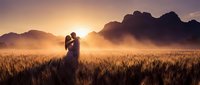 Romantic couple silhouette embracing in golden wheat field at sunset with dramatic mountain backdrop creating dreamy wedding or engagement scene.