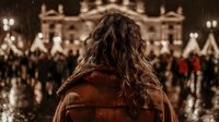 Woman with long curly hair from behind, wearing brown coat, looking at illuminated historic building with crowd at night during rainy evening.