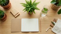 Flat lay of an open kraft notebook on a wooden desk with green plants, pencils, a water glass, and a linen pouch for workspace and lifestyle concepts.