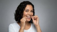 Woman with dark curly hair and medium skin tone flossing teeth against neutral background. Dental hygiene, oral care routine.