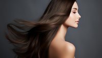 Young Asian woman with flowing brown hair against dark background, profile view showing natural makeup and bare shoulders for beauty and haircare advertising.