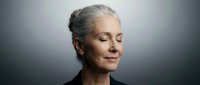 Mature woman with gray hair and fair complexion wearing black clothing, looking downward with serene expression against neutral gray background for portrait use.